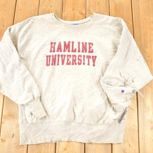 Może przedstawiać: Jasnoszara bluza Hamline University z długimi rękawami. Tekst "HAMLINE UNIVERSITY" jest wydrukowany na piersi na czerwono. Logo Champion jest widoczne na lewym rękawie.