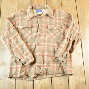 May include: A tan and coral plaid button-down shirt with two chest pockets. The long-sleeved shirt has a collared neckline and buttoned cuffs. The fabric appears slightly wrinkled, giving it a vintage look.