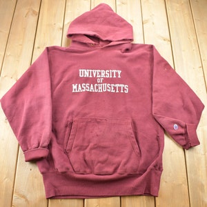 Puede incluir: Una sudadera con capucha Champion de color granate con el texto "University of Massachusetts" en letras blancas. La sudadera tiene un aspecto desgastado y un pequeño logotipo de Champion en la manga izquierda.