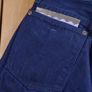 May include: Close-up of a dark blue denim back pocket with a stitched W-shaped design. The pocket has a small metal button and a piece of paper sticking out.