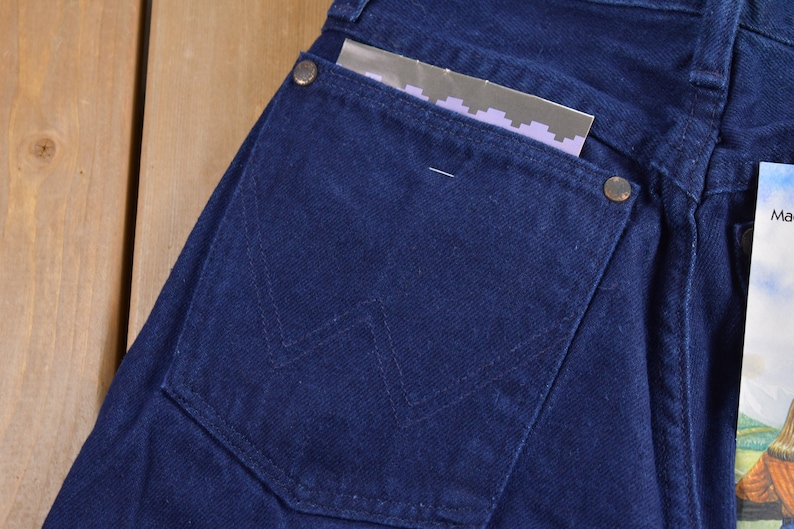 May include: Close-up of a dark blue denim back pocket with a stitched W-shaped design. The pocket has a small metal button and a piece of paper sticking out.