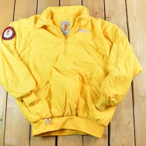 May include: Bright yellow vintage anorak jacket with a half-zip closure, a small collar, and a round patch on the left sleeve. The jacket has a ribbed hem and cuffs. The jacket is made of a lightweight material.