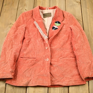 May include: A coral corduroy blazer with a white lace trim and two fabric buttons. The blazer has two pockets and a contrasting fabric collar.