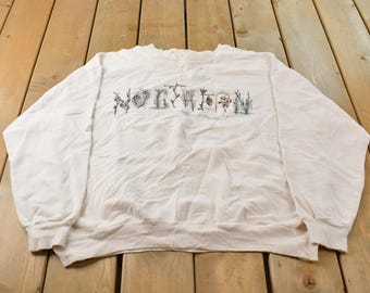 May include: A cream-colored long-sleeve crewneck sweatshirt featuring the word NORTHERN printed across the chest in a dark, stylized font with small, detailed illustrations of pine trees and forest elements integrated into the letters.