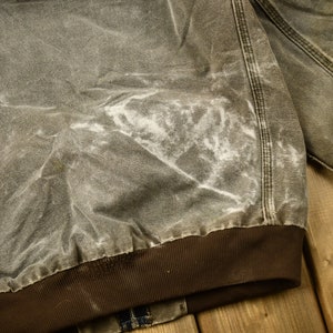 Może przedstawiać: Zbliżenie na brązową kurtkę roboczą z białą plamą z przodu. Kurtka ma brązowy, żebrowany mankiet i jest wykonana z grubego, wytrzymałego materiału.