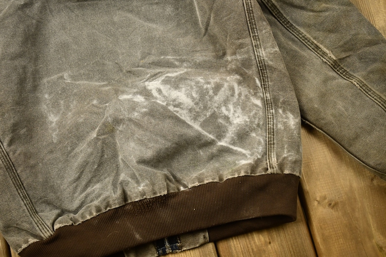 Może przedstawiać: Zbliżenie na brązową kurtkę roboczą z białą plamą z przodu. Kurtka ma brązowy, żebrowany mankiet i jest wykonana z grubego, wytrzymałego materiału.