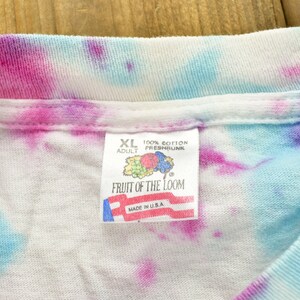 May include: A white tie-dye t-shirt with a Fruit of the Loom tag. The tag is white with red, blue, and green accents. The tag says "XL Adult 100% Cotton Preshrunk Fruit of the Loom Made in U.S.A."