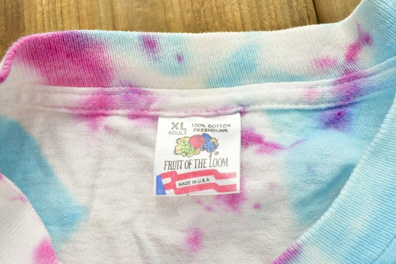 May include: A white tie-dye t-shirt with a Fruit of the Loom tag. The tag is white with red, blue, and green accents. The tag says "XL Adult 100% Cotton Preshrunk Fruit of the Loom Made in U.S.A."