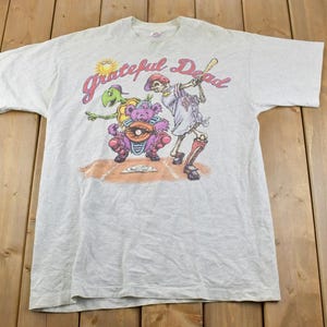 May include: A light grey t-shirt featuring a graphic print of a skeleton, a turtle, and a bear playing baseball. The text "Grateful Dead" is printed above the graphic.