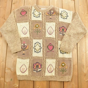 Peut inclure: Un pull en tricot marron avec un motif patchwork composé de différents motifs floraux et de cœurs dans des tons de rose, jaune, vert et bleu.