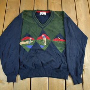 May include: A navy blue V-neck sweater with a green diamond pattern on the chest featuring golf course scenes. The sweater has long sleeves and a ribbed hem and cuffs. The golf scenes include golfers, flags, and water features.