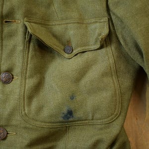 May include: A close-up of a green military jacket with a large pocket on the front. The jacket has several buttons and a flap on the pocket.