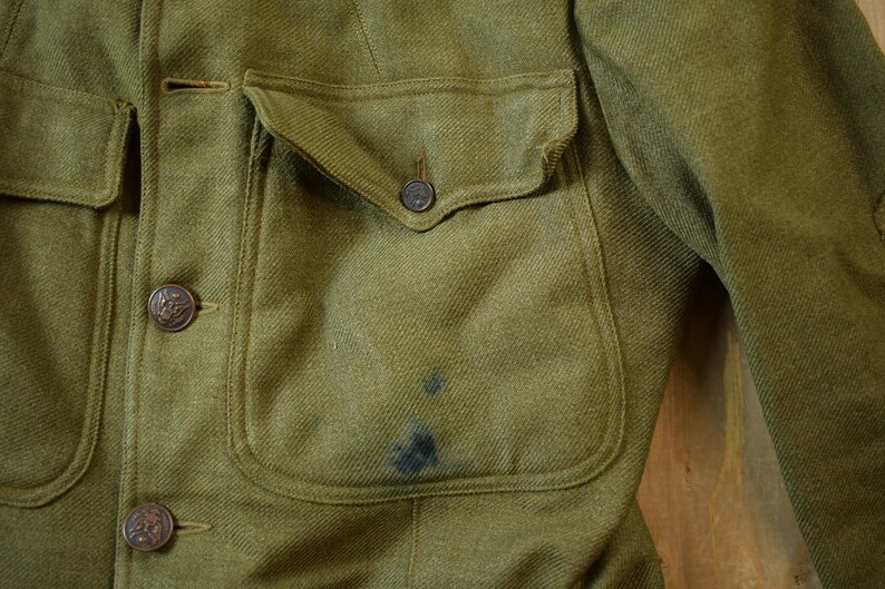 May include: A close-up of a green military jacket with a large pocket on the front. The jacket has several buttons and a flap on the pocket.
