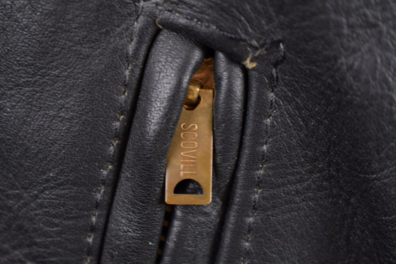 May include: Close-up of a black leather jacket zipper with a gold Scovill zipper pull.