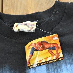 May include: Black crew neck t-shirt with a Spider-Man 2 tag attached. The tag features a graphic of Spider-Man in a dynamic pose. The shirt has a blue gradient design at the bottom.