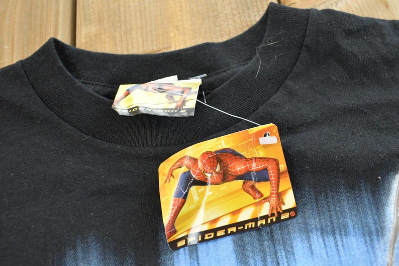 May include: Black crew neck t-shirt with a Spider-Man 2 tag attached. The tag features a graphic of Spider-Man in a dynamic pose. The shirt has a blue gradient design at the bottom.