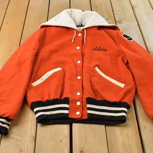 Pode incluir: Uma jaqueta varsity laranja com capuz branco, botões de pressão brancos e dois bolsos frontais com acabamento branco. A jaqueta possui punhos e bainha canelados com listras pretas e brancas, com o nome Adria bordado em preto no peito.