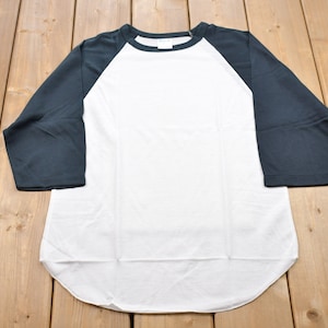 Op de afbeelding: Een wit en marineblauw baseballshirt met driekwart mouwen. Het shirt heeft een witte body en marineblauwe mouwen. Het shirt is gemaakt van een zacht materiaal en ligt op een houten ondergrond.