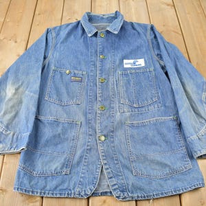 May include: A blue denim work jacket with a patch on the left chest that says "Columbia". The jacket has two large pockets on the front and two smaller pockets on the chest.
