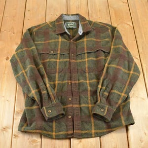 May include: A long-sleeve button-down shirt featuring a plaid pattern in shades of olive green, dark brown, and mustard yellow. The shirt includes a pointed collar, two chest pockets with flaps, and buttoned cuffs. A Woolrich brand label is visible.