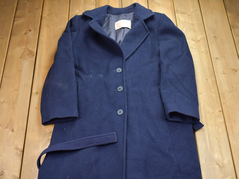 Pu&ograve; includere: Un cappotto di lana blu navy con cintura e quattro bottoni. Il cappotto ha uno stile classico e sartoriale.
