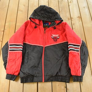 May include: Red and black Chicago Bulls hooded jacket. The jacket has a full zip, black sleeves, and a red body. The Chicago Bulls logo is on the chest. The sleeves have white and black stripes.