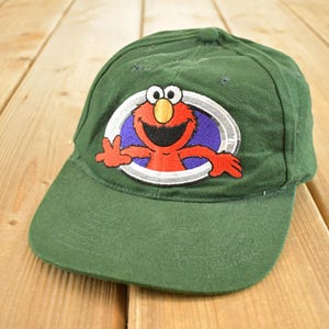 Op de afbeelding: Groene baseballpet met een kleurrijk Elmo-ontwerp. Het Elmo-ontwerp staat in een witte ovaal met een paarse achtergrond. De pet heeft een gebogen klep en een klassiek zes-panelen ontwerp.