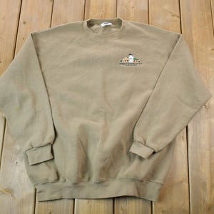 May include: A tan sweatshirt with a small embroidered graphic of a building with the text "The Lighthouse Restaurant" underneath.