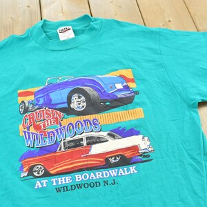 May include: A teal short-sleeve t-shirt featuring a graphic print of two classic cars. The top car is blue, and the bottom car is red and white. The text reads Cruisin the Wildwoods at the Boardwalk Wildwood N.J. in blue and white lettering.