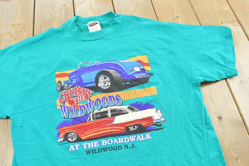 May include: A teal short-sleeve t-shirt featuring a graphic print of two classic cars. The top car is blue, and the bottom car is red and white. The text reads Cruisin the Wildwoods at the Boardwalk Wildwood N.J. in blue and white lettering.