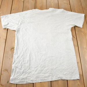 May include: A plain, light grey t-shirt with short sleeves. The t-shirt is laid flat on a wooden surface.
