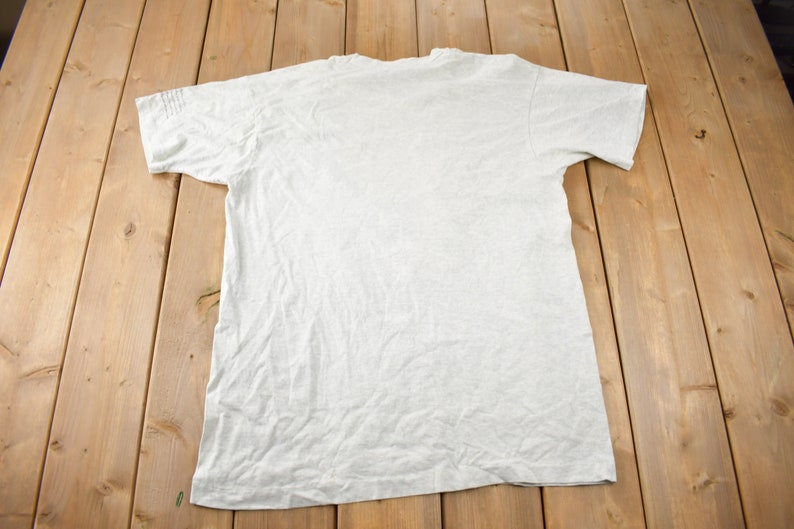 May include: A plain, light grey t-shirt with short sleeves. The t-shirt is laid flat on a wooden surface.