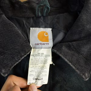 Puede incluir: Primer plano del cuello de una chaqueta Carhartt negra con textura de pana. La etiqueta de la chaqueta presenta el logotipo de Carhartt e informaci&oacute;n del producto. El cuello es oscuro y contrasta con la etiqueta.