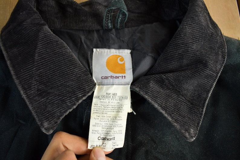Puede incluir: Primer plano del cuello de una chaqueta Carhartt negra con textura de pana. La etiqueta de la chaqueta presenta el logotipo de Carhartt e informaci&oacute;n del producto. El cuello es oscuro y contrasta con la etiqueta.