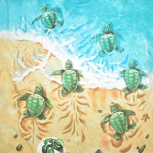 Puede incluir: Una camiseta tie-dye con una escena de playa con seis tortugas marinas beb&eacute;s. Las tortugas son verdes y nadan en el oc&eacute;ano o caminan por la arena. El fondo es un patr&oacute;n tie-dye azul y amarillo.