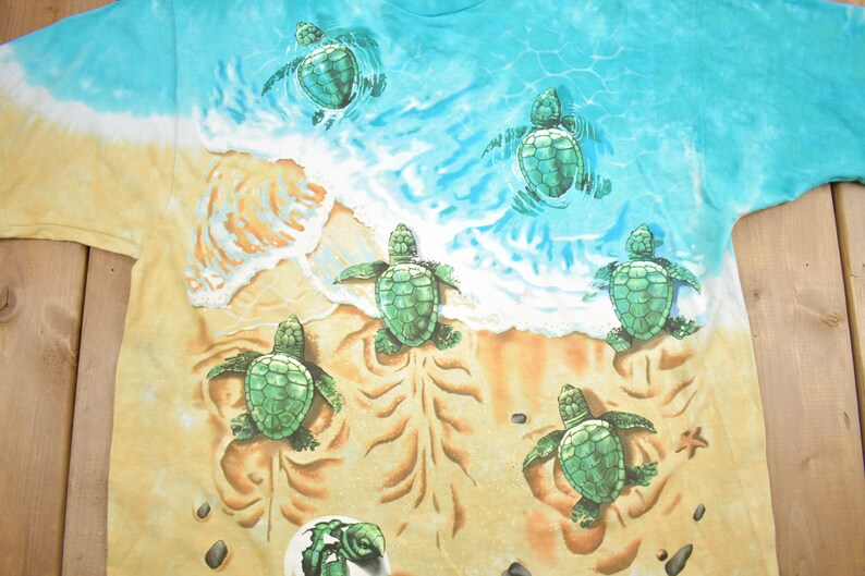 Puede incluir: Una camiseta tie-dye con una escena de playa con seis tortugas marinas beb&eacute;s. Las tortugas son verdes y nadan en el oc&eacute;ano o caminan por la arena. El fondo es un patr&oacute;n tie-dye azul y amarillo.