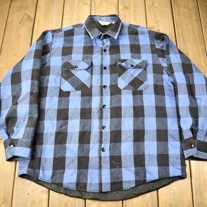 May include: A blue and black checked flannel shirt with long sleeves and a button-up front. The shirt has two chest pockets.