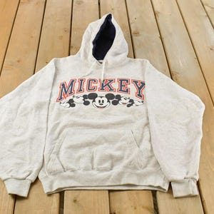 Op de afbeelding: Een lichtgrijs gem&ecirc;leerde hoodie met lange mouwen, een donkerblauwe voering in de capuchon en een kangoeroezak. Op de borst staat het woord MICKEY in marineblauwe blokletters met een rode omlijning boven verschillende Mickey Mouse-gezichten.