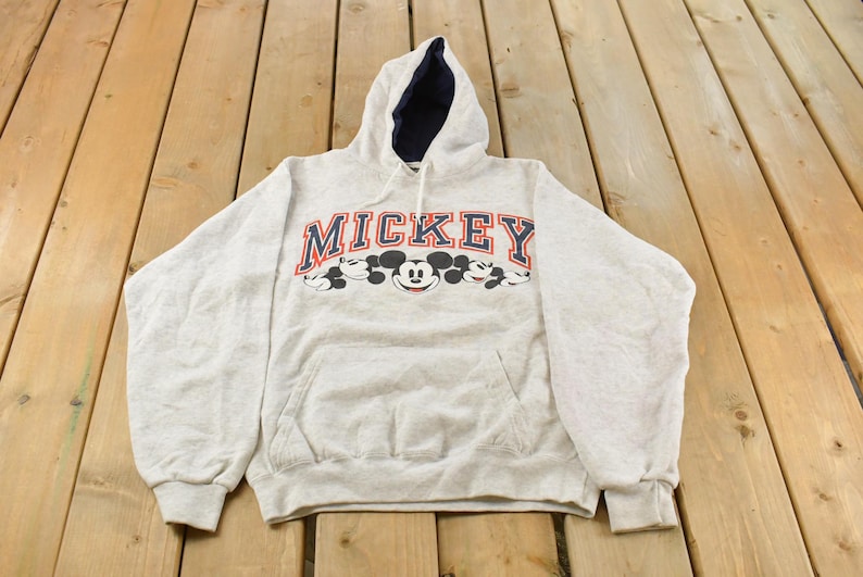 Op de afbeelding: Een lichtgrijs gem&ecirc;leerde hoodie met lange mouwen, een donkerblauwe voering in de capuchon en een kangoeroezak. Op de borst staat het woord MICKEY in marineblauwe blokletters met een rode omlijning boven verschillende Mickey Mouse-gezichten.