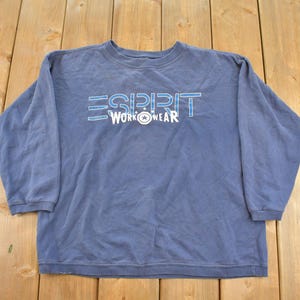 May include: A blue long-sleeve sweatshirt featuring the text ESPRIT in a light blue outline font and the word WORKWEAR in white with a small star graphic centered between the two words on the front of the garment.