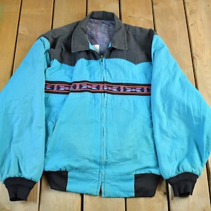 May include: A turquoise jacket with a black collar and cuffs. The jacket features a colourful geometric pattern across the chest. The jacket has a full zip and is made of a lightweight material.