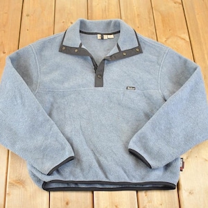 Könnte beinhalten: Hellblaues Fleece-Pullover mit Kragen und Knopfleiste. Das langärmlige Shirt hat schwarze Verzierungen an Kragen, Manschetten und Saum. Ein kleines Logo ist auf der Brust sichtbar.