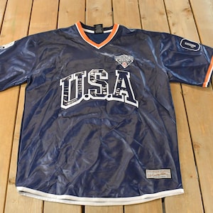 Può includere: Maglia da calcio americana blu navy con scritte e finiture bianche. La maglia ha uno scollo a V con finiture arancioni e un piccolo logo sul petto. Le maniche hanno accenti arancioni e bianchi.