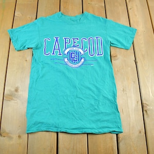 Può includere: Una maglietta turchese con la scritta "CAPE COD" in lettere bianche e blu. Sotto, un emblema circolare con le parole "CAPE COD" e "MASSACHUSETTS" in blu. La maglietta è realizzata in materiale morbido.