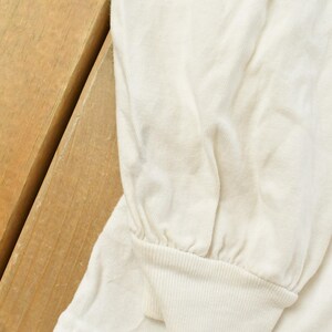 May include: Close-up of a cream-colored long-sleeved shirt. The sleeve is gathered at the wrist with a ribbed cuff. The shirt is made of a soft, slightly wrinkled fabric. The background is a wooden surface.