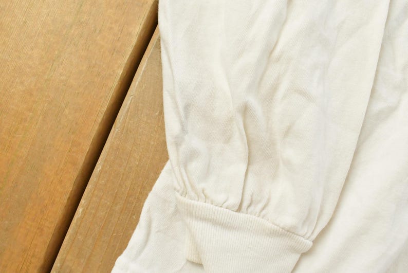 May include: Close-up of a cream-colored long-sleeved shirt. The sleeve is gathered at the wrist with a ribbed cuff. The shirt is made of a soft, slightly wrinkled fabric. The background is a wooden surface.