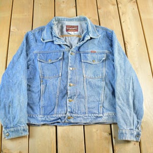 May include: Light blue denim jacket with a button-up front, two chest pockets, and two side pockets. The jacket has a classic collar and buttoned cuffs. The Wrangler brand label is visible on the chest pocket.