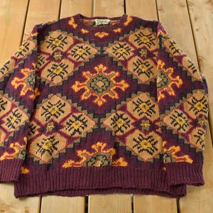 Puede incluir: Un suéter vintage tejido en tonos burdeos, beige, amarillo y negro. El suéter presenta un patrón geométrico con motivos de diamantes y flores. El suéter de cuello redondo se exhibe sobre una superficie de madera.