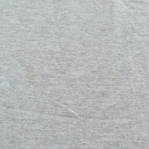 Puede incluir: Primer plano de una tela gris claro con una superficie texturizada. La tela parece ser un material de punto o jersey, con sutiles variaciones de color y textura. La imagen muestra los detalles del textil.