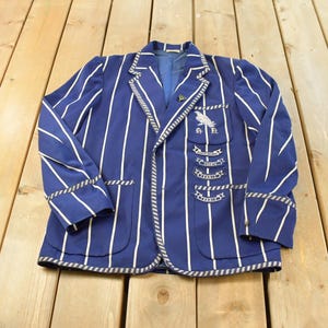 Puede incluir: Un blazer azul con rayas verticales blancas, con solapa de muesca, dos bolsillos delanteros y un bolsillo en el pecho con un emblema de pájaro bordado sobre cuatro parches en forma de cinta con texto, acabado con un borde a rayas.